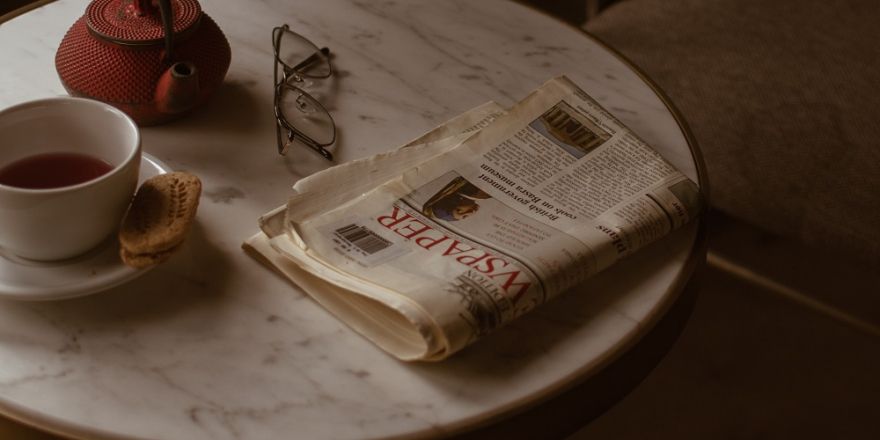 Een tafel met daarop een krant, een (lees)bril, rood theekannetje en een kop thee