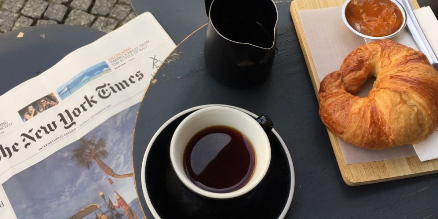 Foto van een tafel met daarbovenop een opengeslagen krant, een kop koffie, een kannetje en een croissant met jam.