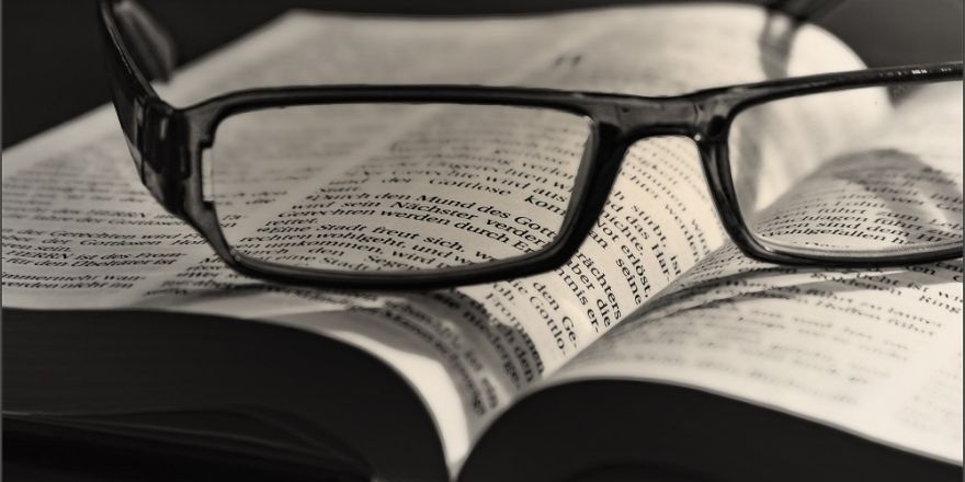 Foto van een opengeslagen boek met daarbovenop een (lees)bril.