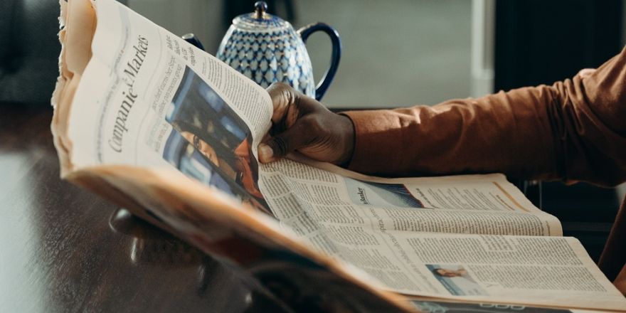 Man houdt krant vast, er staat een theepot naast