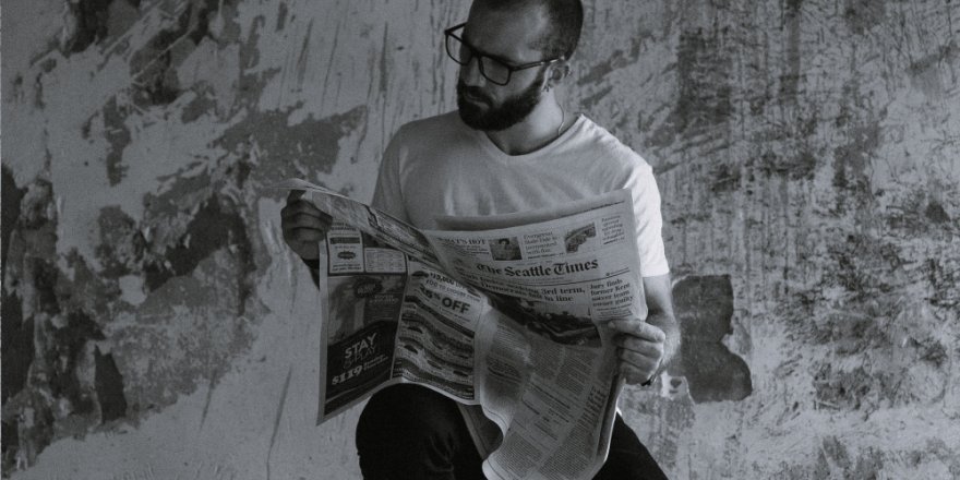 Afbeelding in zwart-wit van een man met een baard en een bril die op een kruk een krant leest