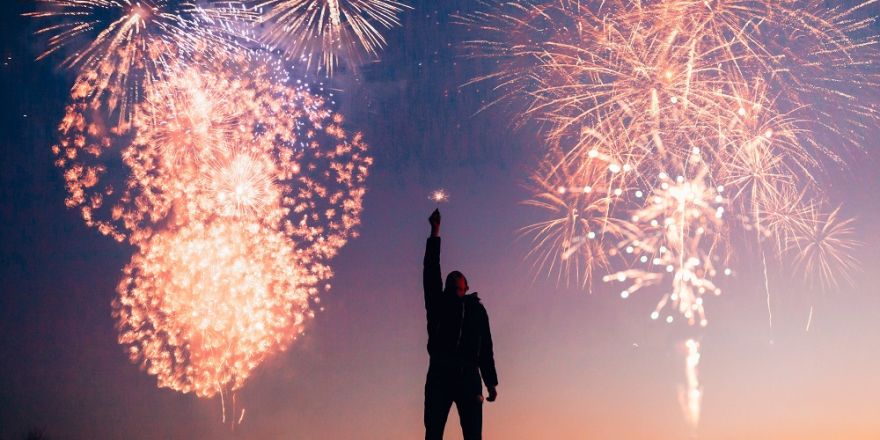 Een persoon met een vlammetje in zijn hand, met op de achtergrond een zonsondergang en vuurwerk