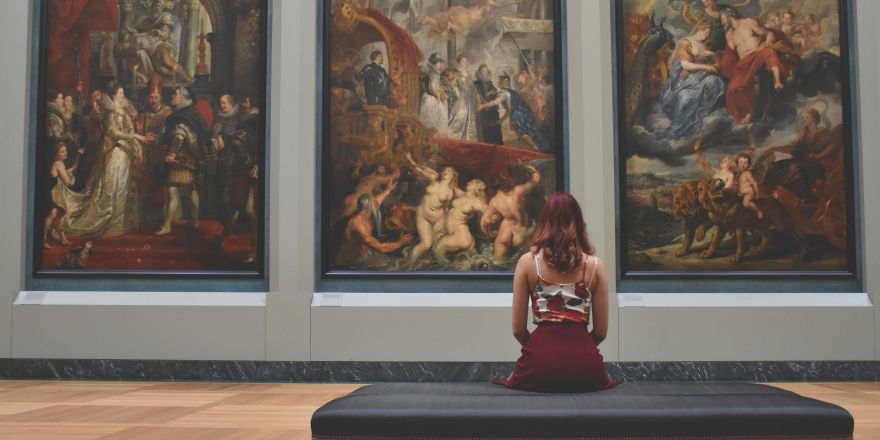 3 schilderijen aan de muur in een museum, vrouw zit met rug op bankje naar schilderijen te kijken