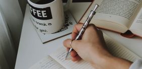 Afbeelding van (de hand van) iemand die aan het schrijven is in een schrift, op de foto staan ook nog een kop koffie en een opengeslagen boek.
