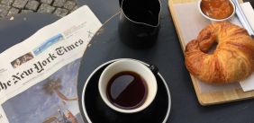 Foto van een tafel met daarbovenop een opengeslagen krant, een kop koffie, een kannetje en een croissant met jam.