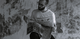 Afbeelding in zwart-wit van een man met een baard en een bril die op een kruk een krant leest