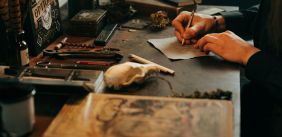 Persoon die iets schrijft op een oud blaadje, om diegene heen liggen boeken en schrijfgerei. 